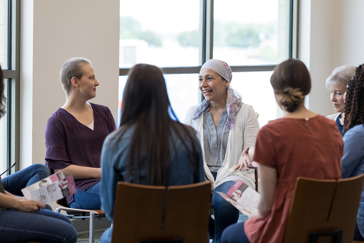 Group of cancer survivors sit in a support group circle and listen to a smiling member speak.
